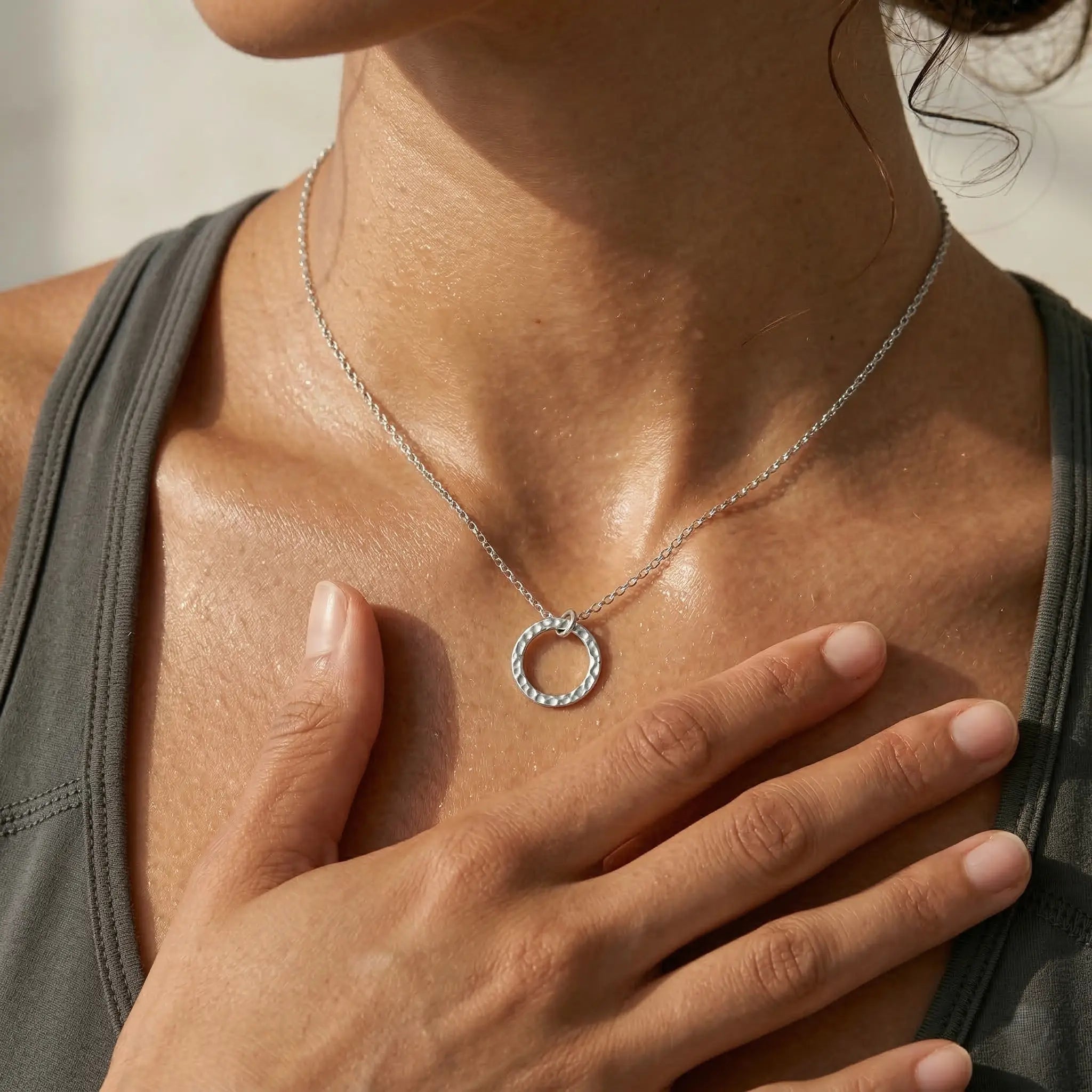 Woman wearing a silver necklace with a circular pendant, hand on chest.